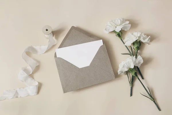 Matrimonio femminile, composizione di articoli di cancelleria di compleanno. Biglietto d'auguri vuoto, mockup d'invito con busta artigianale. Fiori di garofano bianco e nastro di seta su sfondo tavolo beige. Posa piatta, vista dall'alto. — Foto Stock