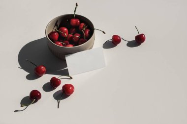 Modern yiyecekler hala yaşam kompozisyonu. Bej masa arkasında güneş ışığında kırmızı kiraz seramik kasesi. Boş kartvizit sahnesi. Üst Manzara. Sert uzun gölgeli yaz meyvesi kompozisyonu.