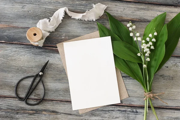 Spring wedding stationery. Blank greeting card, invitation mockup. Black vintage scissors, silk ribbon and bouquet of blooming lilly of the valley flowers on old wooden background. Flat lay, top view