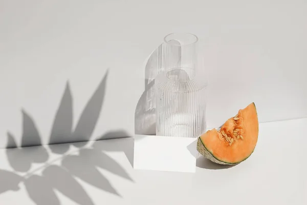 Composición de naturaleza muerta de comida de verano. Melón melón en la mesa. Pared blanca con cubierta de sombra de hoja de palma. Tarjeta de papel en blanco, maqueta de invitación. Jarra de cristal de agua, cóctel a la luz del sol. — Foto de Stock