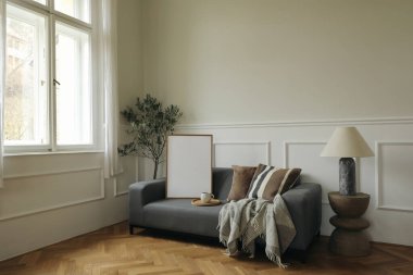 Modern minimalist oturma odası içi gri kanepe, ahşap zemin, dekoratif yastıklar, rahat battaniye, mermer lamba, geniş pencereye yakın ev bitkisi. İskandinav tarafsız ev tasarımı. Resim çerçeve modeli