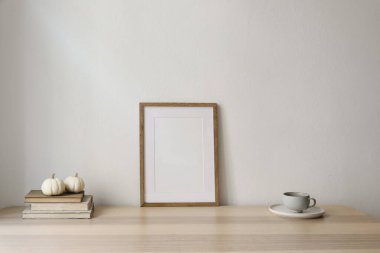 Zarif bir iç mekan. Kış sanat kompozisyonu. Masada boş tahta çerçeve poster modeli var. Bej duvar arka planı. Bir fincan kahve, çay. Beyaz balkabakları, kitaplar, Şükran Günü, Noel dekorasyonu.