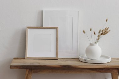 Minimalist ev dekorasyonu. İki boş çerçeve. Tahta bank, masa. Kurumuş lagurus çiçekli beyaz seramik vazo. Sanat eserleri, baskılar, posterler için modern nötr iç modeller. Damgalama.