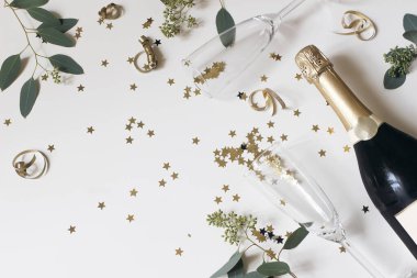 Yeni yılınız kutlu olsun. Boş şampanya bardakları, şarap şişesi. Altın yıldız konfetisi, okaliptüs ağacı dalları beyaz masa arkasında izole edilmiş. Noel kutlaması, parti. Düz üst görünüm.