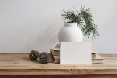 Noel hala hayattır. Boş tebrik kartı davetiyesi maketi, eski kitaplar. Ahşap masa bankında bulanık çam ağacı dalları olan seramik vazo. Kağıt Noel süsleri, toplar, şablon sahnesi.