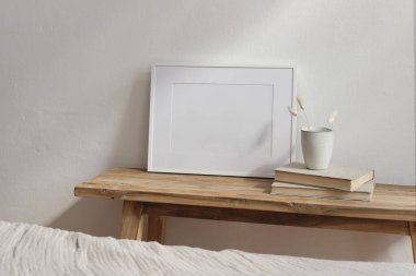 Yatay beyaz resim çerçevesi poster modeli. Antika ahşap masa, seramik bardaklı bank, tavşan kuyruğu kuru çimenler, eski kitaplar. Beyaz duvar arka planı. Bulanık yatak önü, İskandinav ev içi.