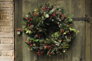 Eski püskü ahşap kapıya asılı Noel çelengi. Bulanık tuğla duvar arka planı. Yeşil köknar ağacı dalları, okaliptüs, kırmızı böğürtlen ve sarmaşıktan oluşan tatil çiçekleri. Kış çiçekleri, yapraklar.