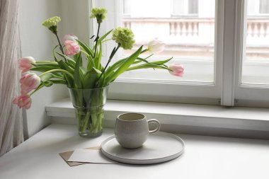 Paskalya baharı hala hayat. Bir fincan çay kahve ve çiçek buketi pencere eşiğinde. Pembe laleler, vazoda yeşil karanfil çiçekleri. Boş tebrik kartı maketi. Ev dekorasyonu, kahvaltı, İskandinav iç mimarisi.