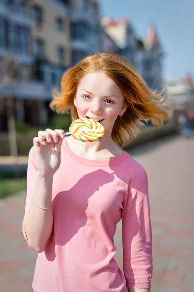 Redhead beautiful young woman biting a lollipop. Pretty girl having fun outdoors. — Stockfoto