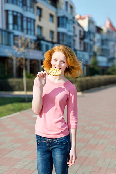 Wanita muda berambut merah cantik menggigit permen lolipop. Gadis cantik bersenang-senang di luar ruangan . — Stok Foto