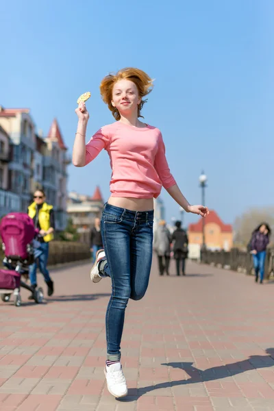 Redhead beautiful young woman jumping high in air over blue sky holding colorful lollipop. Pretty girl having fun outdoors. — Stock Photo, Image