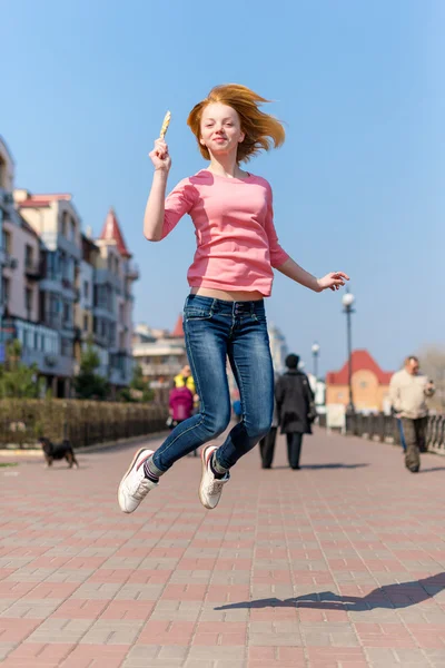 Redhead beautiful young woman jumping high in air over blue sky holding colorful lollipop. Pretty girl having fun outdoors. — Φωτογραφία Αρχείου