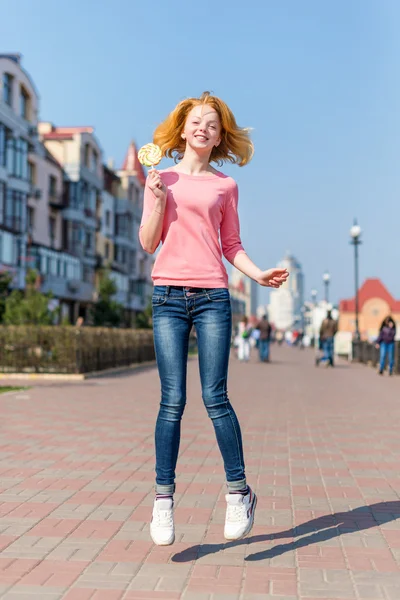 Redhead beautiful young woman jumping high in air over blue sky holding colorful lollipop. Pretty girl having fun outdoors. — Stock Photo, Image
