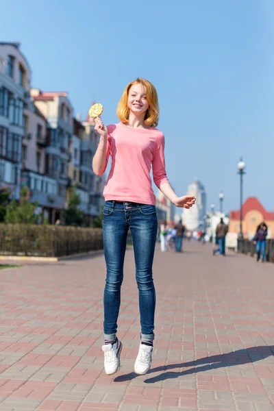 Redhead beautiful young woman jumping high in air over blue sky holding colorful lollipop. Pretty girl having fun outdoors. — Stock Photo, Image