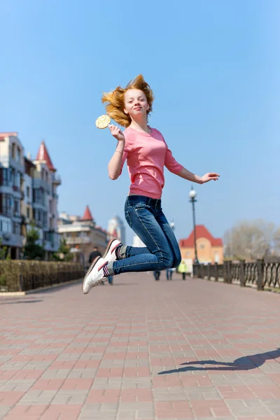 Redhead beautiful young woman jumping high in air over blue sky holding colorful lollipop. Pretty girl having fun outdoors. — Stock Photo, Image