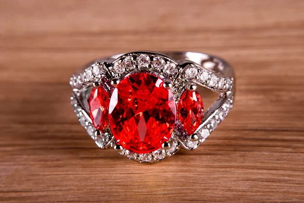 Macro view of silver or white gold ring with red gem and diamonds on wooden background.