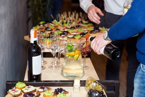 Waiter pouring red wine in a glass at a restaurant table full of appetizers with guests standing near.