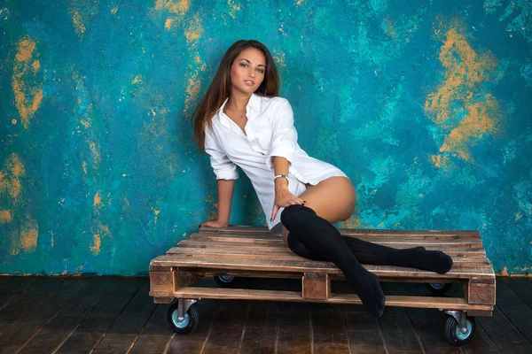 Sexy young woman in a mans shirt and leg warmers sitting on wooden pallet — Stock Photo, Image
