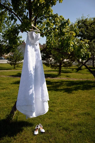 Vestido de noiva pendurado em uma árvore no parque e sapatos femininos brancos — Fotografia de Stock