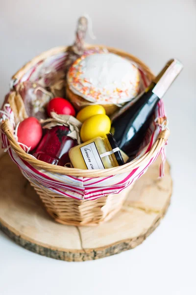 Ovos de páscoa coloridos em uma cesta com bolo, vinho tinto, hamon ou salsicha fumada seca e seca em fundo branco . — Fotografia de Stock