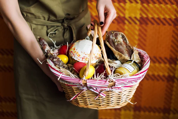 Женские руки держат корзину с разноцветными яйцами, тортом, красным вином, гамоном или вяленой и сухой копченой колбасой. Набор для празднования Пасхи . — стоковое фото