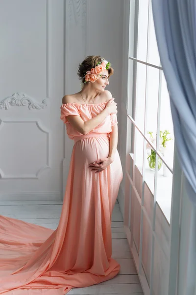 Mulher grávida em vestido de noite rosa em pé perto da janela. Tiro de moda . — Fotografia de Stock