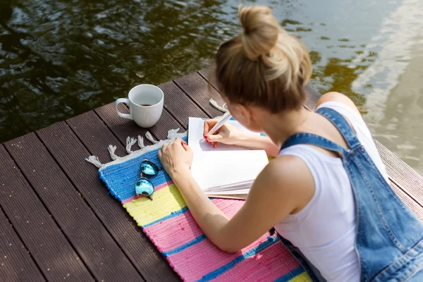 Woman making plans, taking notes in calendar or writing in her diary lying on the pier and drinking hot coffee. Daydreaming, planning concept — Stock Photo, Image