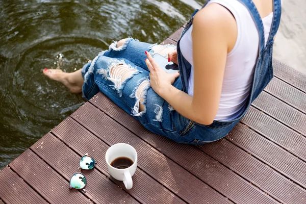 Mujer joven usando su teléfono inteligente sentado en el muelle y bebiendo café caliente — Foto de Stock
