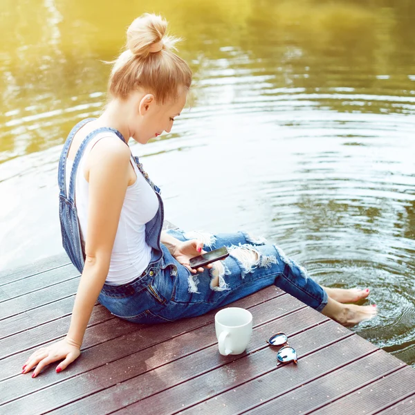 Молодая женщина с помощью смартфона сидит на пирсе и пьет горячий кофе — стоковое фото