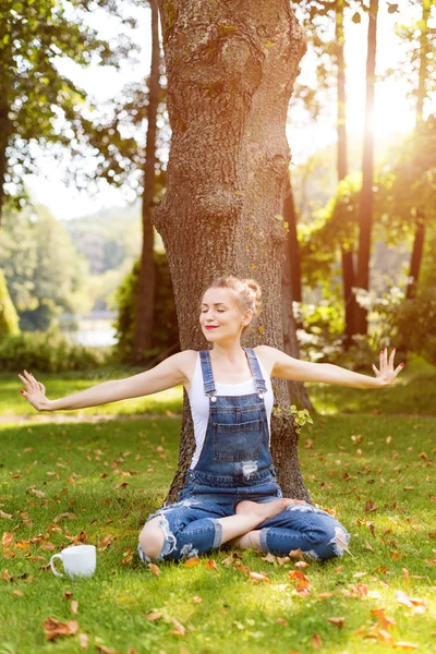 Young blonde woman drinking coffee or tea in the forest sitting in lotus pose and enjoying the warm weather in autumn. — Stock Photo, Image