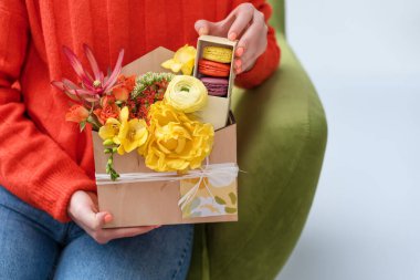 Elinde çiçekli ve şekerli hediye kutusu olan bir kadın. Tahta kutuda makarna ve bahar buketi.