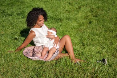 Young african american woman breastfeeding her baby sitting on grass in summer park. Infancy, motherhood, lactation, nutrition and outdoors breastfeeding concept. Black mother breast feeding her child clipart