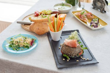 Healthy burger with hamon, tomatoes, micro greens and black wholegrain buns, vegetable sticks on the table. Cooking with microgreens. Clean eating, dieting, detox food concept