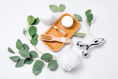 Masaj silindiri ve fırça, temel yağ veya kozmetik serumu ve beyaz arka planda okaliptüs yapraklı kozmetik kremi. Güzellik ürünleri, masaj aletleri, yüz bakımı, güzellik tedavi konsepti.