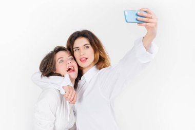 İki kadın ışık stüdyosunda selfie çekerken poz veriyor. Kızların en iyi arkadaşı kamerada komik suratlar yapıyor, dil gösteriyor ve birlikte gülüyorlar.