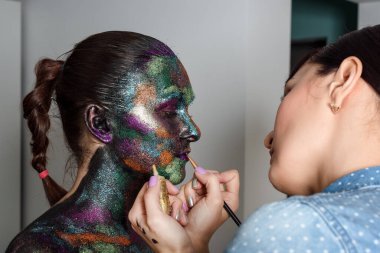 Makyöz, siyah boya ve renkli pudrayı genç modele uyguluyor. Uzay ve yıldızlar teması üzerine yaratıcı vücut sanatı. Vücut boyama projesi: sanat, güzellik, moda