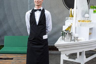 Açık büfe yemek partisinde şarap servisi yapan üniformalı profesyonel garsonun orta bölümü, şenlik ya da düğün. Tepside şampanya dolu bardaklar. Açık hava partisi yemek servisi, garsonluk.