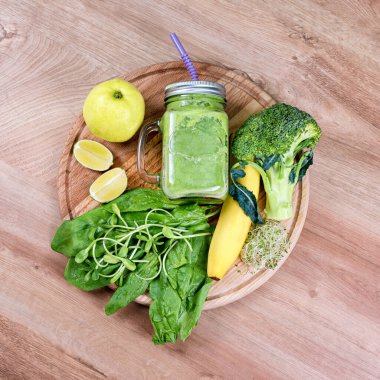 Taze yeşil sebze ve kavanozda yeşil meyve püresi. Detoks, diyet ya da sağlıklı gıda konsepti. Tahta zemin üzerinde brokoli, ıspanak, mikroyeşillik, limon ve muz bulunan bir kavanoz diyet içeceği. Üst görünüm