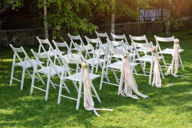 Güzel bir düğün hazırlığı. Bahçede düğün seremonisi. Çiçeklerle süslenmiş beyaz ahşap sandalyeler ve sırada duran pembe kurdeleler.