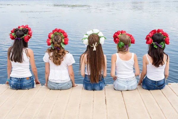 Back view of five young ladies, wearing flower wreaths, jeans and white tshirts. Group of friends sitting on the side of the pool or on pier. Pool party, summer vacation. Banner for website