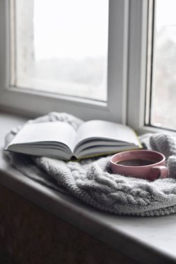 Sıcak ev hayatı: bir fincan sıcak kahve ve dışarıdaki kar manzarasına karşı pencerenin kenarında sıcak ekoseli kitap. Kış tatili ve Noel zamanı konsepti, bedava fotokopi alanı.