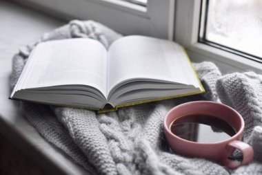 Sıcak ev hayatı: bir fincan sıcak kahve ve dışarıdaki kar manzarasına karşı pencerenin kenarında sıcak ekoseli kitap. Kış tatili ve Noel zamanı konsepti, bedava fotokopi alanı.