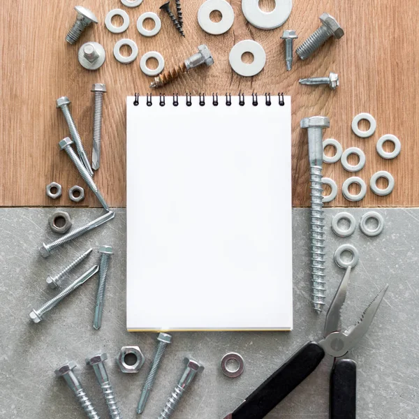Construction tools. The screws, nuts and bolts arranged around blank spiral bound note book paper on wooden and concrete background. Repair, home improvement concept. Free space for text, top view, flat lay