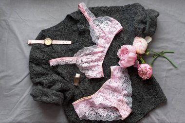 Modaya uygun kadın iç çamaşırı manzarası. Şakayıklar, kızların güzellik ürünleri, parfüm. Güzel pembe dantelli kadın iç çamaşırı. İç çamaşırı ve sutyen gri örgü süveter arka planda yatıyor.