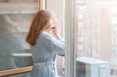 Bornozlu sarışın kadın sabah kahvesini ya da çayını şehir merkezindeki bir balkonda içiyor. Gün doğumunu ve şehir merkezini balkondan seyreden bir kadın.