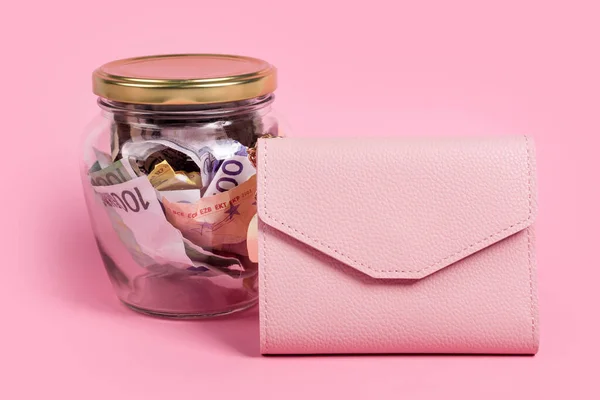 Euro banknotes in glass money jar with blank label and pink wallet, financial, saving. Money box with empty sticky note paper. Jar full of cash, save money concept, expense planning and control.