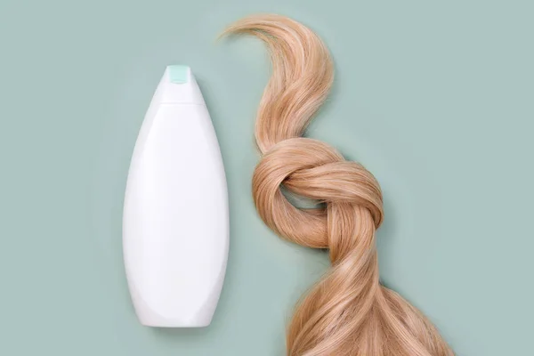 Shampoo or conditioner bottle and blonde hair lock tied in knot. Strand of honey blonde hair, top view. Hairdresser service, hair strength, haircut, hairstyle, hair extension, treatment concept