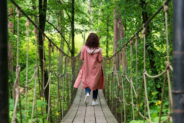 Young woman walking on suspension bridge in the forest. Traveler on the rope bridge. Travel woman on vacation concept. Hiking, summer outdoors activity.