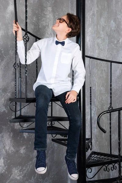Розумний підліток носить окуляри, сидячи на спіральних сходах в квартирі лофт. Дівчина-підліток у білій сорочці позує в лофт — стокове фото