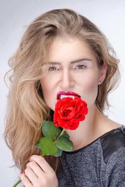 Linda chica rubia con colmillos de vampiro y maquillaje brillante mordiendo una flor —  Fotos de Stock